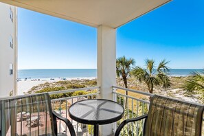 Un balcon avec une table et des chaises en verre, donnant sur une plage bordée de palmiers et surplombant un ciel bleu azur.