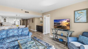 A modern living room with a sofa, coffee table, and TV. A kitchen is visible in the background. - Beach Front Corner Unit - Beach Place #212 (Madeira Beach)