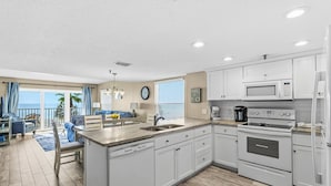 A modern kitchen with white cabinets, a built-in microwave, and an oven. There is a dining area with a table and chairs, and a view of the ocean through large windows. - Beach Front Corner Unit - Beach Place #212 (Madeira Beach)