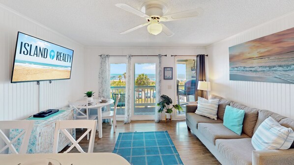 A room with a ceiling fan, a TV displaying 'ISLAND PENNY,' a dining area with a table and chairs, a sofa with cushions, and a view of the beach through the door.