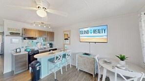 Une cuisine moderne avec un îlot central, un coin repas avec des chaises blanches et un téléviseur diffusant le logo de « ISLAND REALTY ».