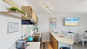 Une cuisine moderne équipée d’une cuisinière intégrée, d’une machine à café et d’un grille-pain. On y trouve également un coin repas avec une table et des chaises, ainsi qu’un téléviseur qui diffuse le programme « ISLAND REALTY ».