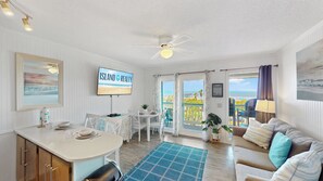 Una stanza con una TV che mostra 'ISLAND REALTY', un'area pranzo con tavolo e sedie, un divano, una ventola a soffitto e vista sulla spiaggia attraverso la porta.
