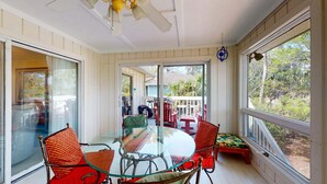 Une pièce lumineuse, baignée de soleil, avec une table et des chaises en verre, de grandes fenêtres et un ventilateur de plafond.