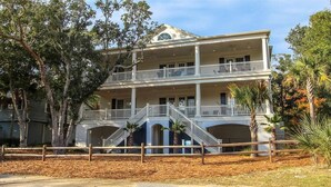 A two-story building with a balcony on the upper floor, surrounded by trees and a fence. - Beautiful beach home with wrapping porches, private pool, and ocean view peek (Isle of Palms)