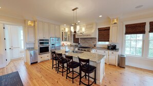 A spacious kitchen with white cabinets, a granite countertop, a central island with bar stools, stainless steel appliances, and a large window with blinds. - Beautiful beach home with wrapping porches, private pool, and ocean view peek (Isle of Palms)