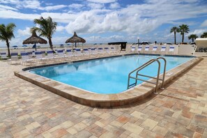 A swimming pool with a tiled deck, lounge chairs, palm trees, and a beach view.
