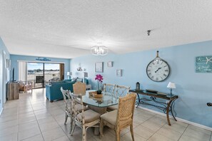 A spacious living area with a glass dining table, wicker chairs, a blue sofa, and a view of the beach through large windows.