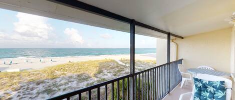 A balcony with a view of a beach and ocean. There is a table and two chairs.
