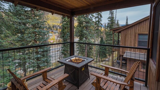 A wooden deck with a stone fire pit and two wooden chairs, overlooking a forested area with a cabin in the background. - Largest Black Bear Townhome - Ping Pong Table - on Demand Ski Shuttle (Durango)