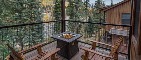 A wooden deck with a stone fire pit and two wooden chairs, overlooking a forested area with a cabin in the background.