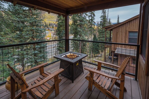Une terrasse en bois avec un foyer en pierre et deux chaises en bois, donnant sur une zone boisée où l’on aperçoit un chalet au loin.