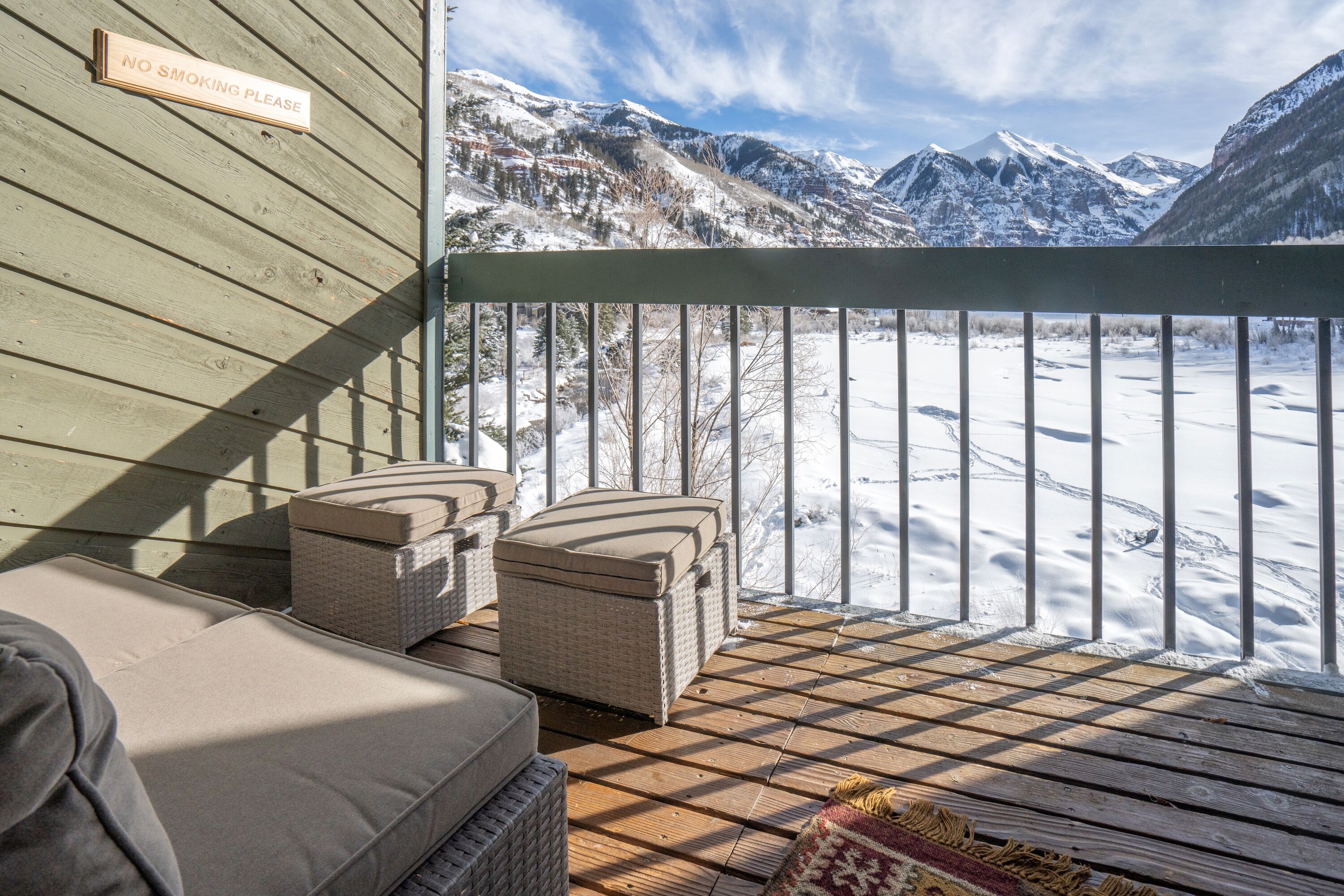 A wooden deck with outdoor furniture, a 'NO SMOKING PLEASE' sign, and a view of snow-covered mountains.