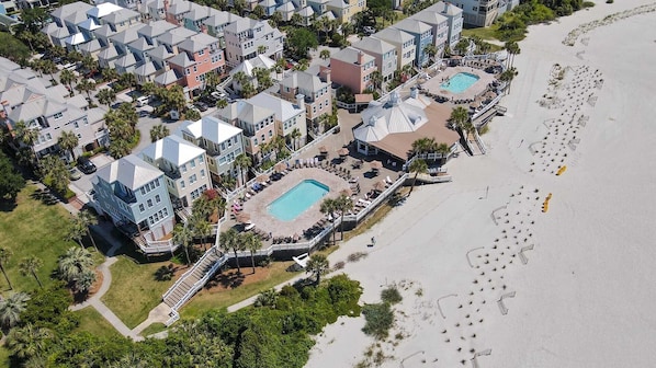 An aerial view of a coastal resort with multiple hotel buildings, a central pool area, and a beach with footprints.