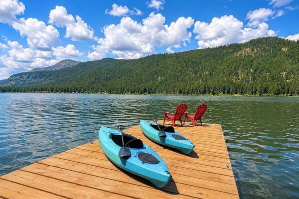 Une jetée en bois, avec des kayaks et des chaises rouges, au bord d’un lac paisible, le tout entouré d’une montagne boisée.