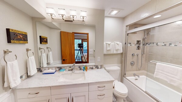 A modern bathroom with a large mirror, a sink, a toilet, and a bathtub. There are towels hanging on hooks and a camera on a tripod in the background.