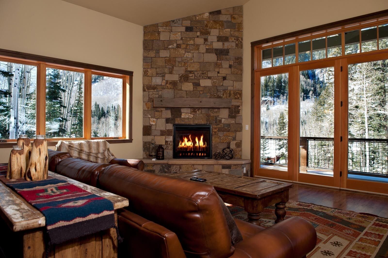 A cozy living room with a stone fireplace, leather furniture, and large windows offering a view of trees.