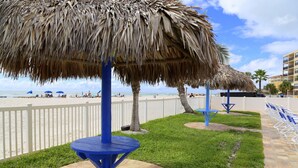 Zona de playa con un paraguas de paja, mesas azules y una cerca blanca.