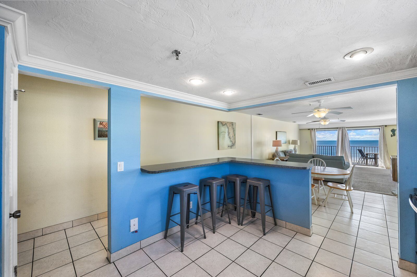 Habitación con zona de cocina que incluye una encimera azul y taburetes, comedor con mesa y sillas, y vista al océano a través de grandes ventanales.