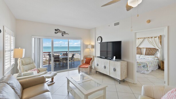 A spacious living room with a television, a sofa, a coffee table, and a balcony with a view of the ocean.