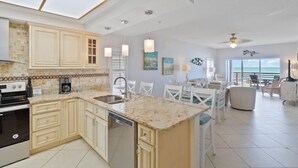 A spacious kitchen with a large island, a dishwasher, and a coffee maker. There is a dining area with a table and chairs, and a view of the ocean through large windows.