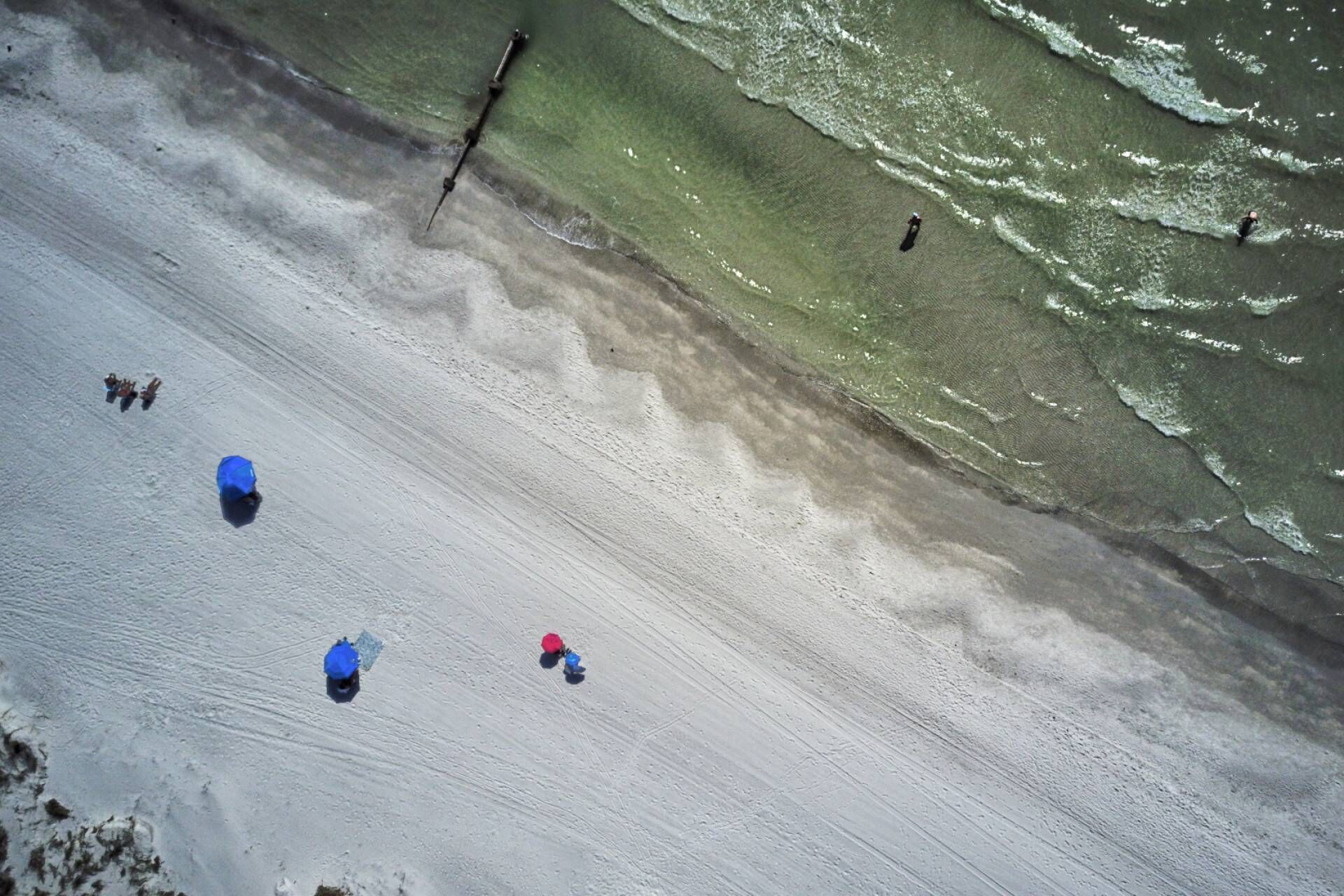 Vue aérienne d’une plage avec une jetée qui s’avance dans l’eau. On aperçoit plusieurs personnes dispersées le long du rivage, certaines près du bord de l’eau et d’autres plus en retrait.