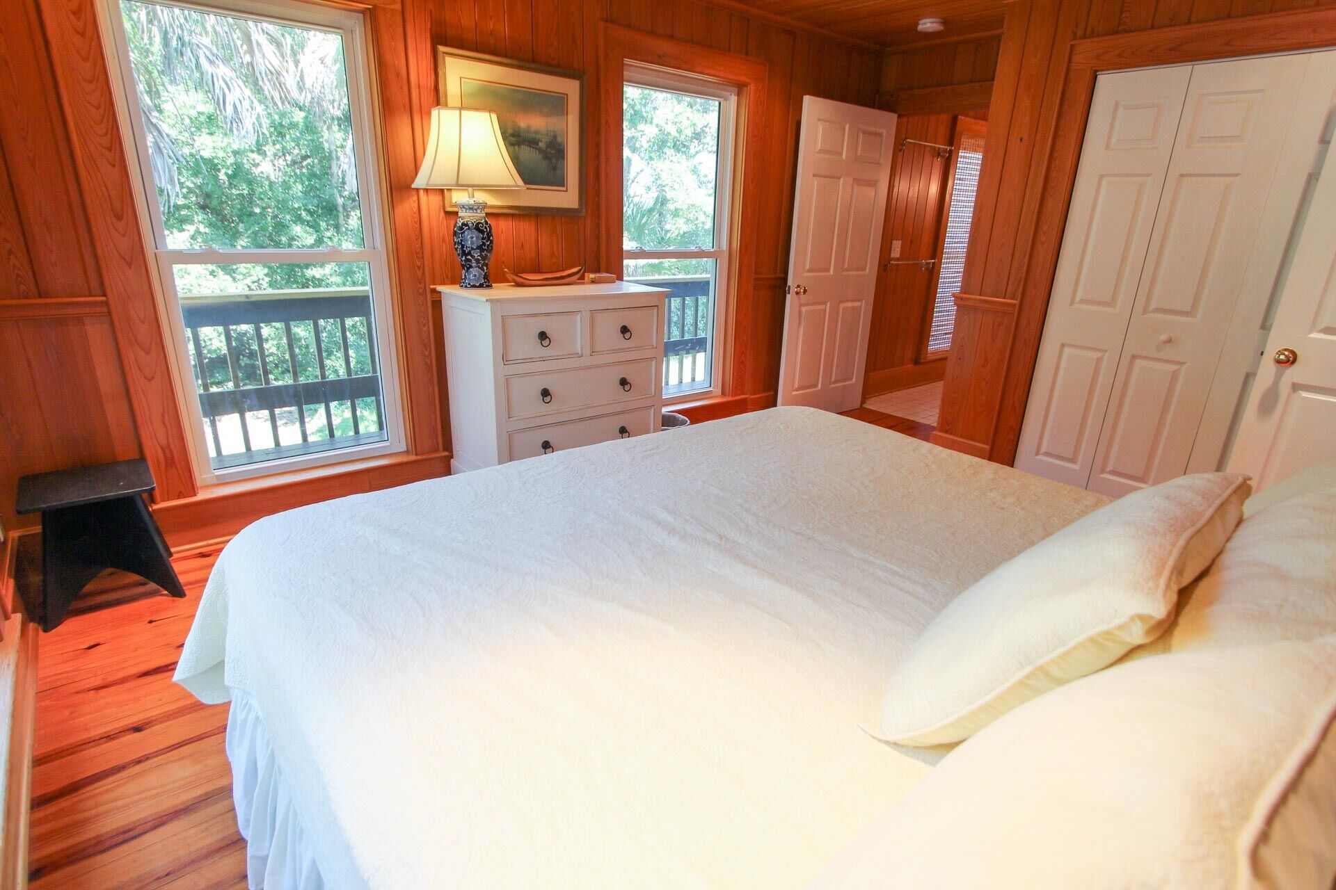 Une chambre à coucher avec un grand lit, une commode, une lampe et une fenêtre donnant sur les arbres.