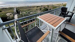 Un balcon avec une table et des chaises en bois, donnant sur une plage où l’on peut voir des gens et des parasols.