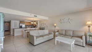 Amplia sala de estar con un sofá color beige, una mesa de centro de vidrio y un comedor con una mesa y sillas blancas. Se ve una cocina al fondo con gabinetes de madera y un candelabro.