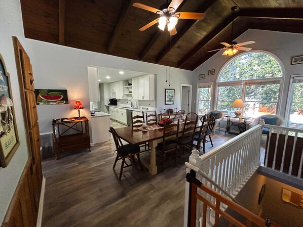 A spacious dining area with a wooden table and chairs, a kitchen in the background, and a view through large windows.