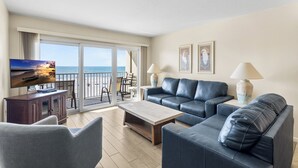 Una sala de estar con sofá, sillón, mesa de centro, televisor y un balcón con vista al mar.