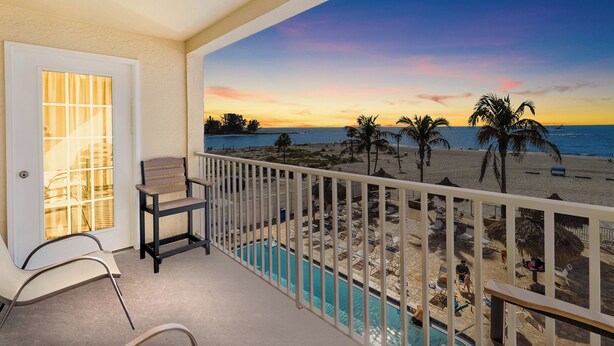 Un balcón con piscina, una puerta y vista a una playa con palmeras y un atardecer.