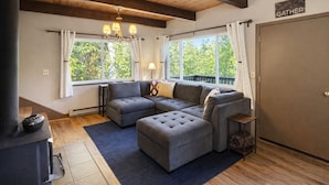 A cozy living room with a sectional sofa, a fireplace, and large windows offering a view of greenery. - Fish Lake | Dock | Hot tub | Dogs | View | Game Rm (Leavenworth)