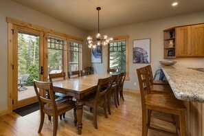A dining area with a wooden table and chairs, large windows, and a chandelier.