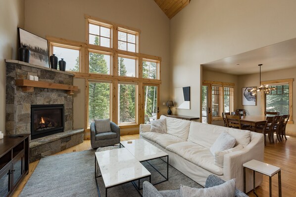 A spacious living room with a stone fireplace, large windows, a white sofa, and a dining area.