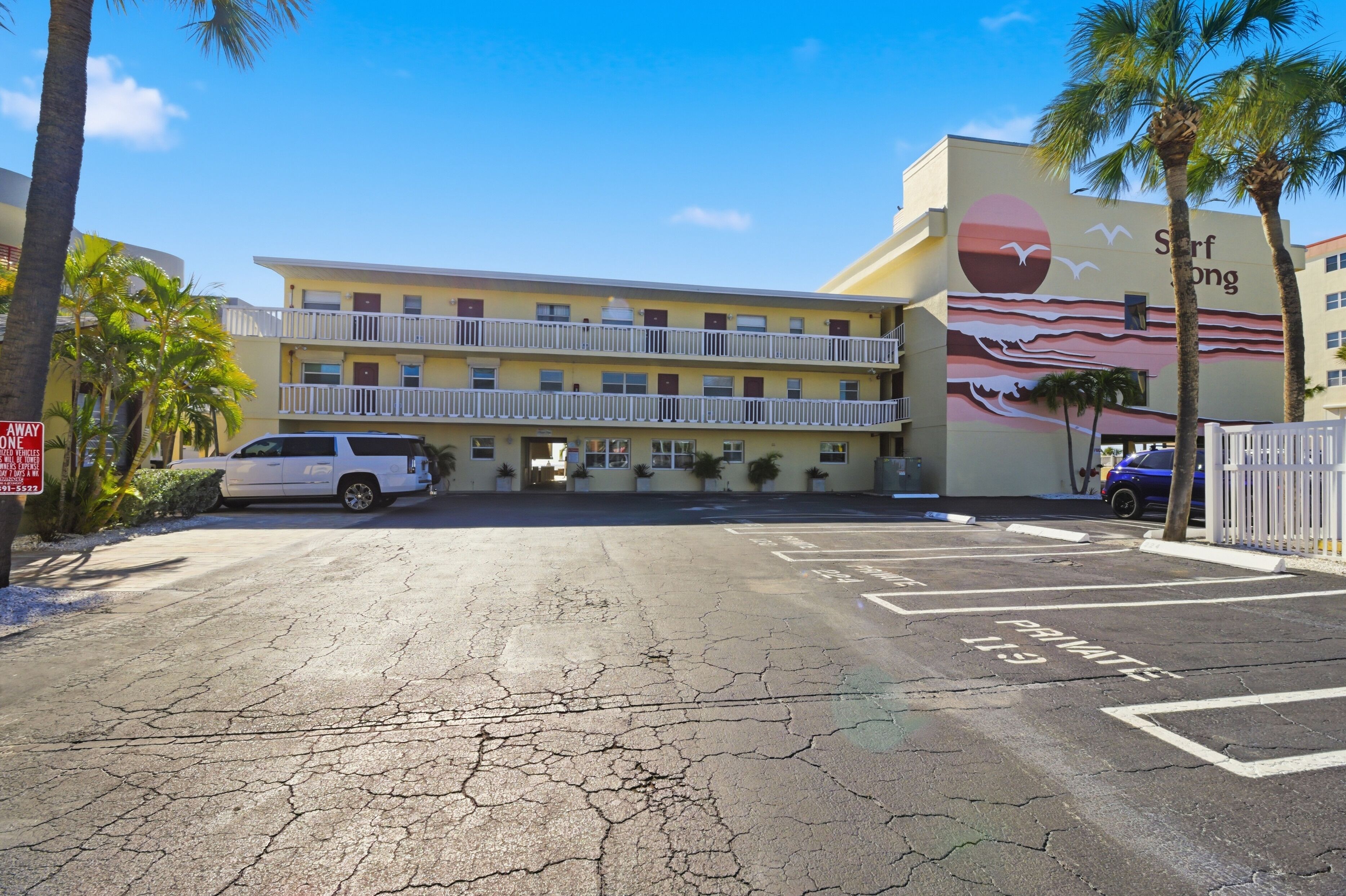 A multi-story hotel with a balcony on the second floor, a parking lot with marked spaces, and a mural on the building's side.