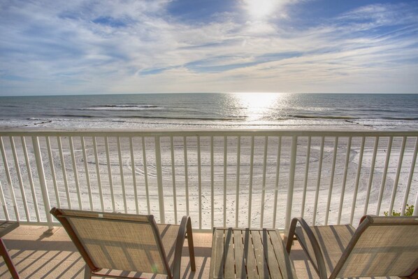 A beach view from a balcony with a railing, featuring two wooden chairs and a small table.