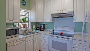 Une cuisine avec des meubles blancs, un four à micro-ondes, un évier, une cuisinière et une horloge au mur.