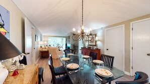Uma sala de jantar com uma mesa de vidro, cadeiras e um candelabro. Há um sofá e uma mesa de jantar ao fundo.
