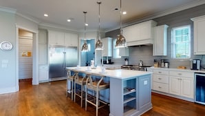 Una cocina moderna con armarios blancos, una isla central con taburetes de bar, electrodomésticos de acero inoxidable, iluminación colgante y una ventana con persianas.