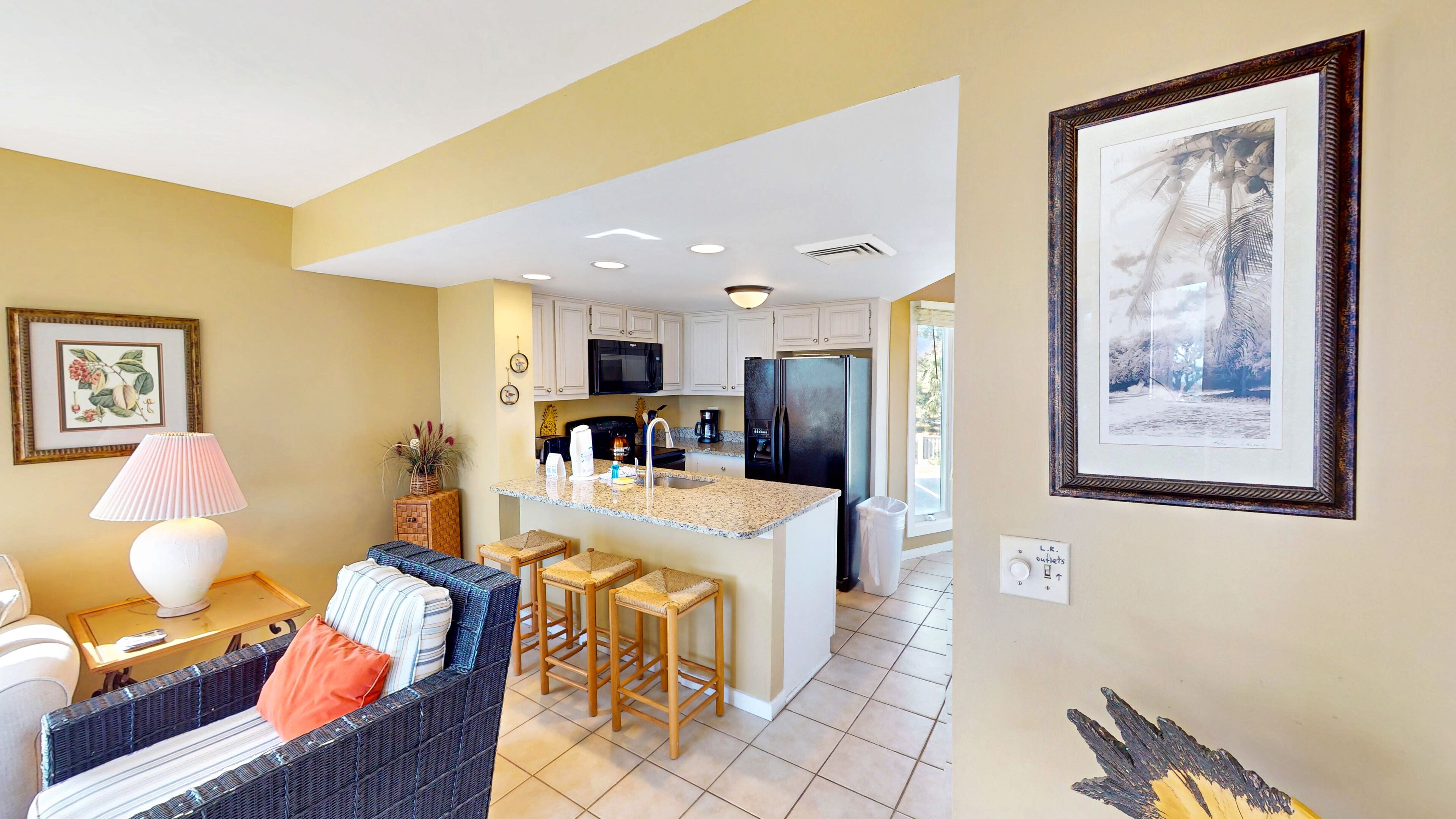 A modern kitchen with white cabinets, a black refrigerator, and a granite countertop. There is a dining area with wicker furniture and a lamp.