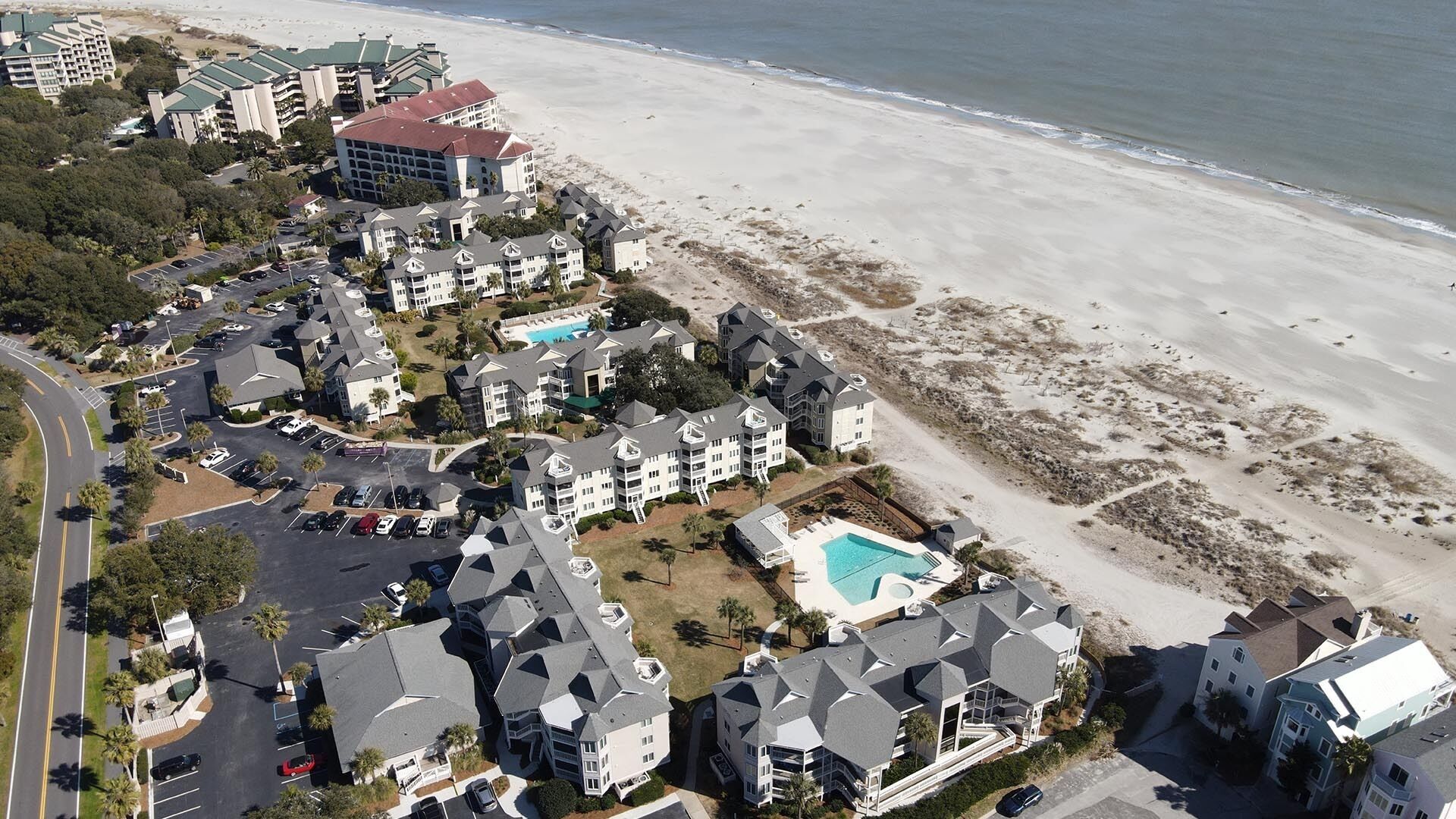 An aerial view of a coastal residential area with multiple two-story houses, a swimming pool, and a beach in the background.