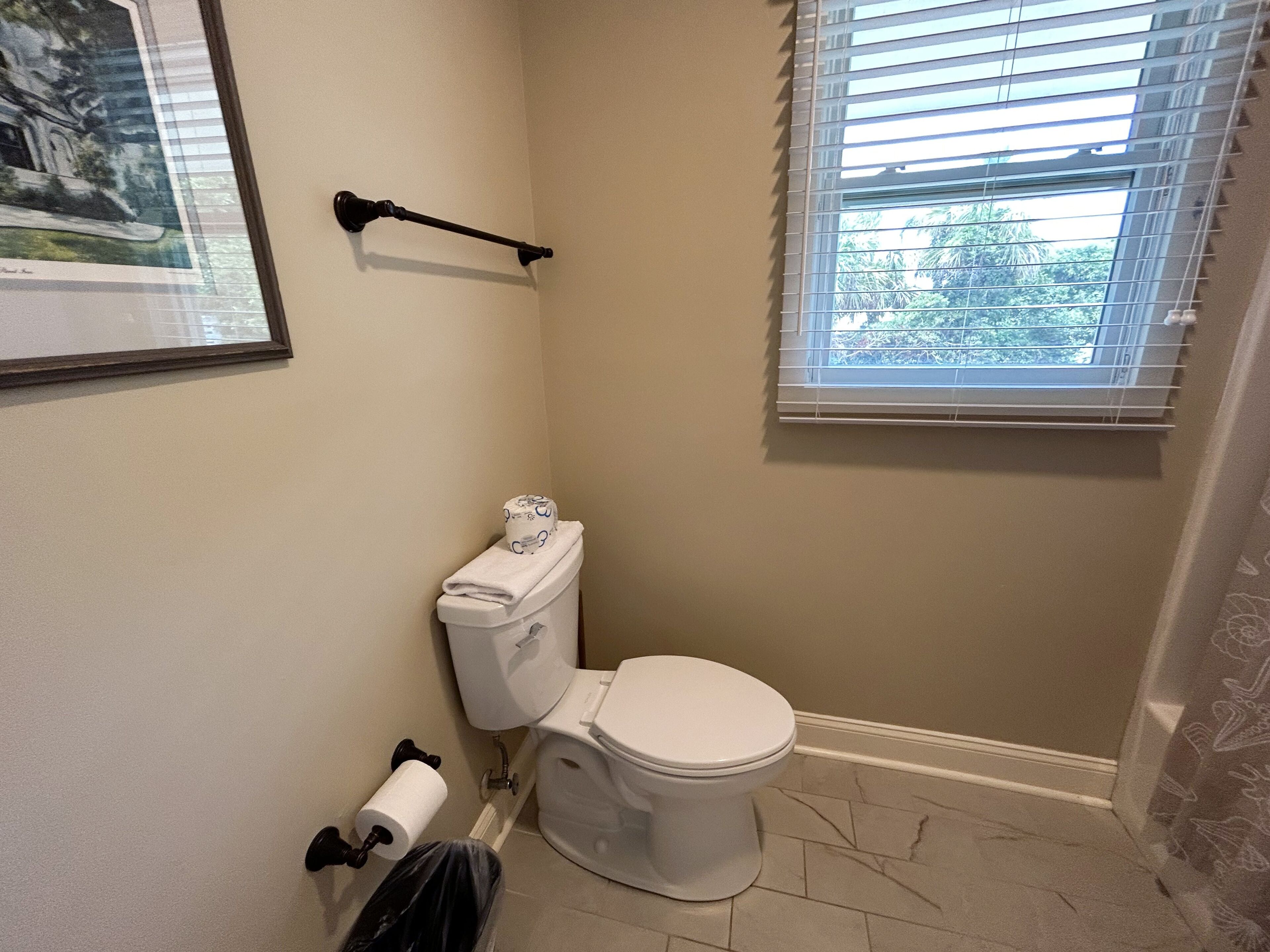 A bathroom with a toilet, a towel rack, a window with blinds, and a framed picture on the wall.