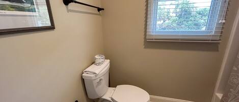 A bathroom with a toilet, a towel rack, a window with blinds, and a framed picture on the wall.