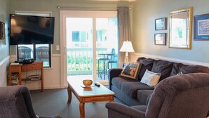 A living room with a flat-screen TV, a sofa, a coffee table, and a view of the outside balcony. - Cozy Condo Steps From the Water! (Isle of Palms)