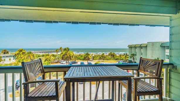 A balcony with two wooden chairs and a table, overlooking a beach and ocean. - Oceanfront Condo with Private Balcony & Outstanding Ocean Views (Isle of Palms)