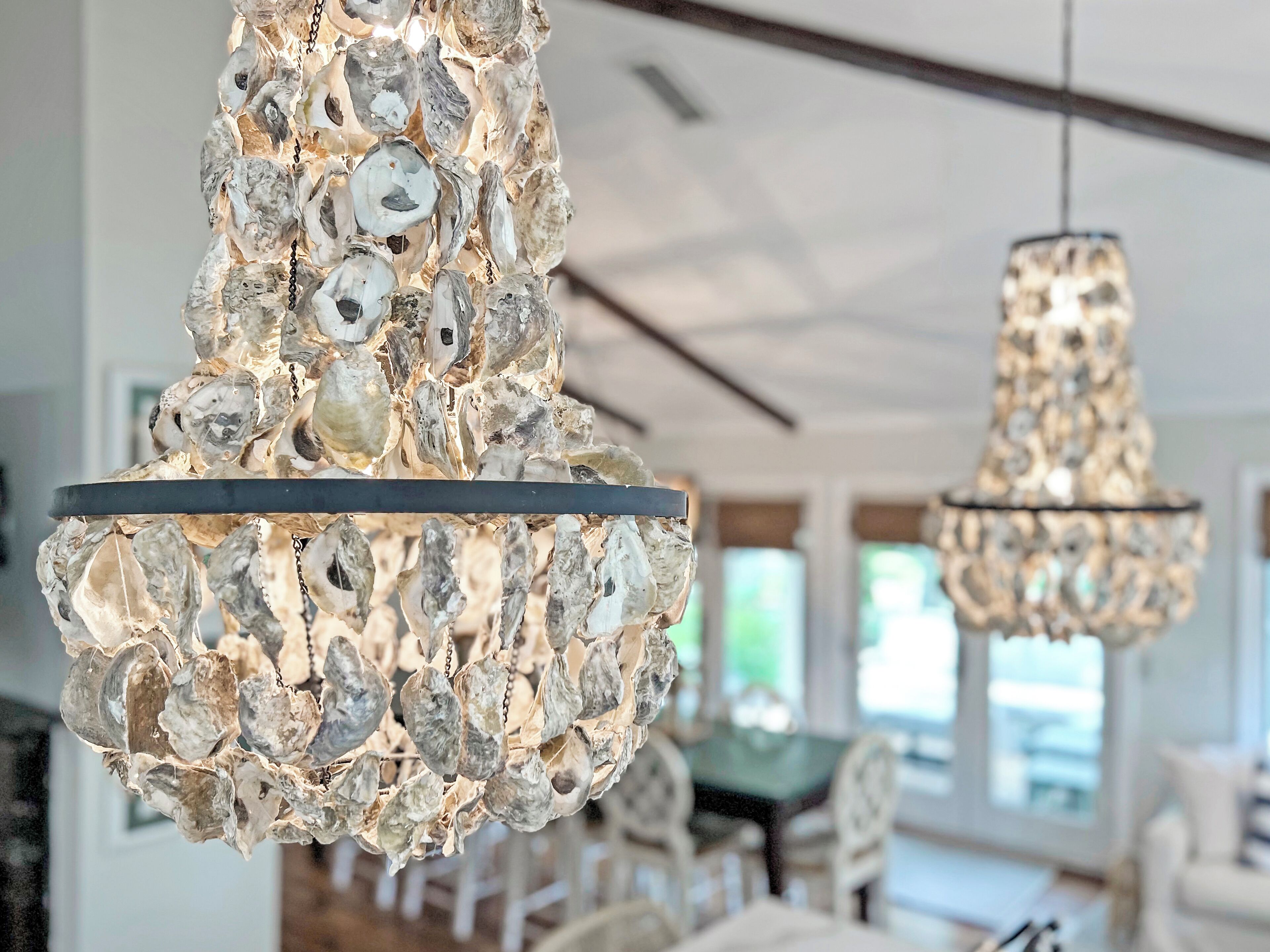 Een kroonluchter van natuurlijke materialen, vermoedelijk schelpen, hangt in een goed verlichte kamer met een eethoek op de achtergrond.