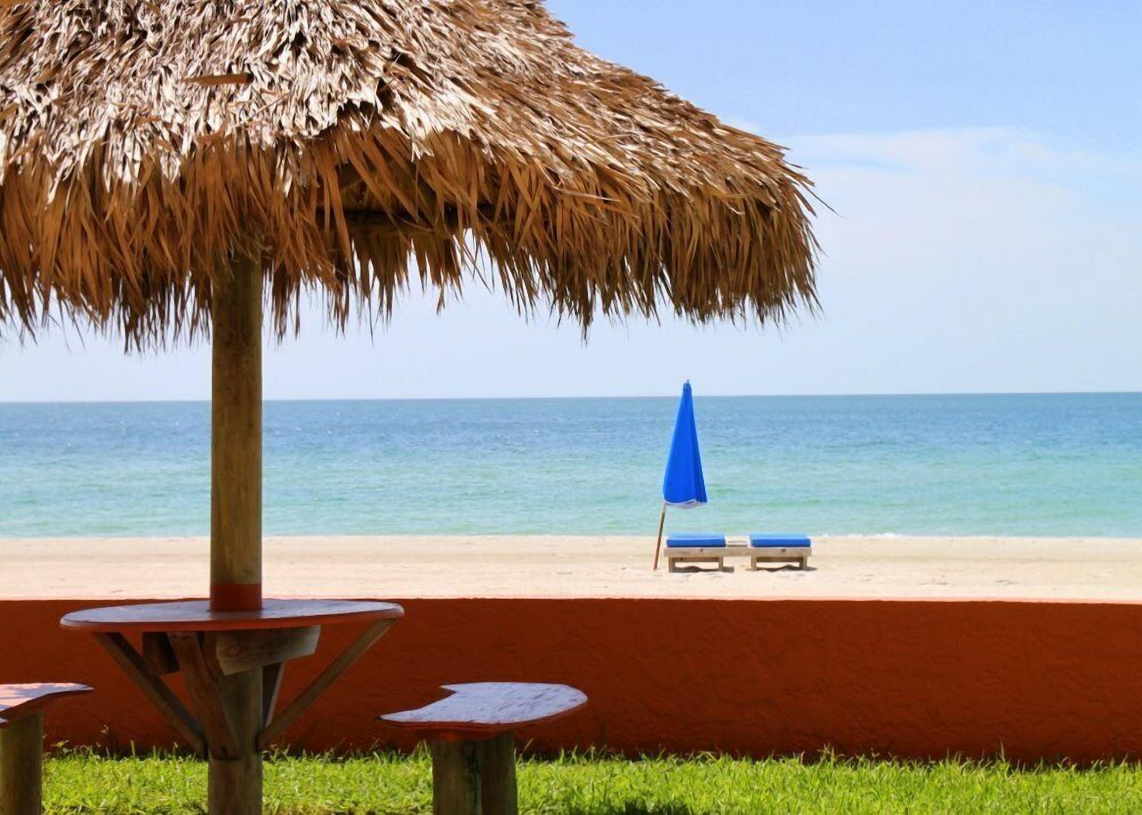 A beach with a thatched umbrella, two blue lounge chairs, and a table.
