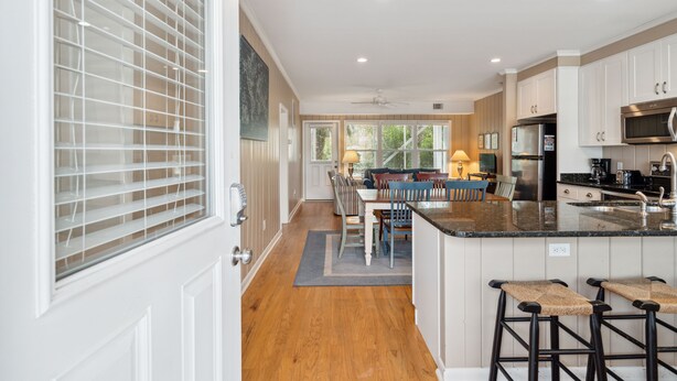 A modern kitchen with white cabinets, a central island with stools, a dining area with a table and chairs, and a view of the outdoors through large windows. - Walk to beach, screened porch, single level, updated kitchen and baths! (Isle of Palms)