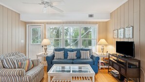A living room with a blue sofa, striped armchair, and a coffee table. There is a television on a wooden stand and a ceiling fan. - Walk to beach, screened porch, single level, updated kitchen and baths! (Isle of Palms)